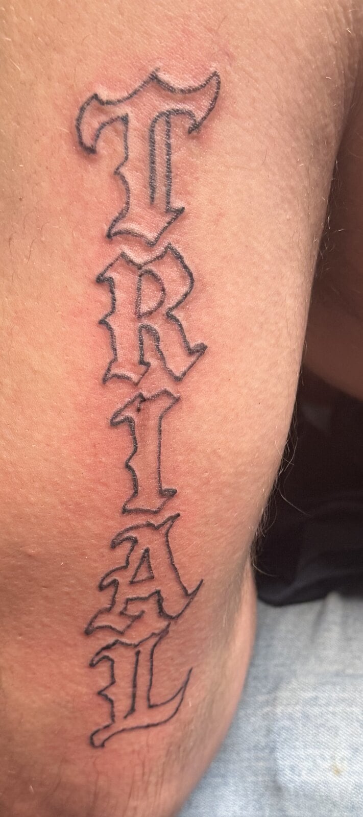 The word TRIAL stacked vertically in outlined blackletter down the back of an arm